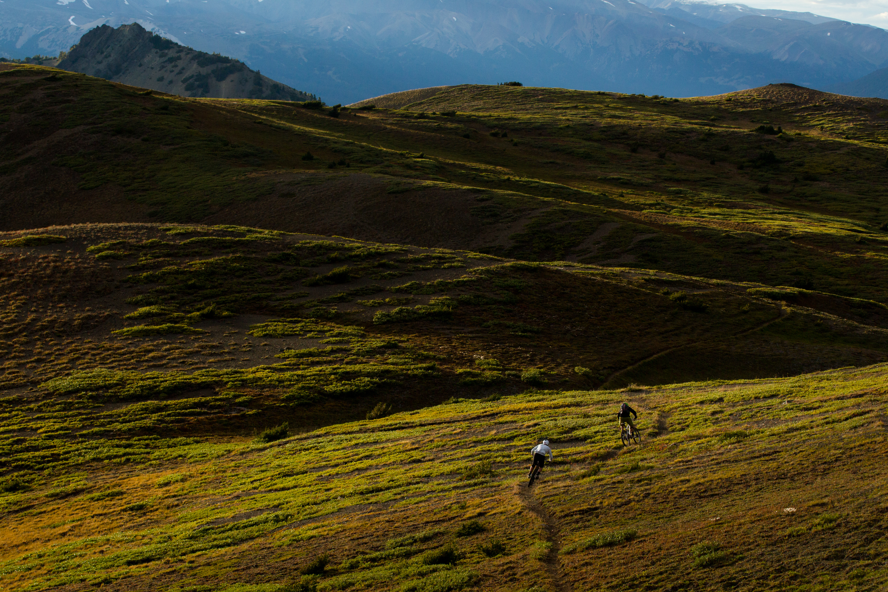 Two riders descending high, sweeping ridgeline grass with peaks behind